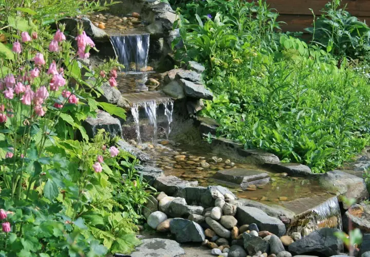Ein kleiner, mehrstufiger Wasserfall im Garten, umgeben von üppigem Grün und rosa Blüten. Tolle wasserfall garten ideen für eine entspannende Oase.