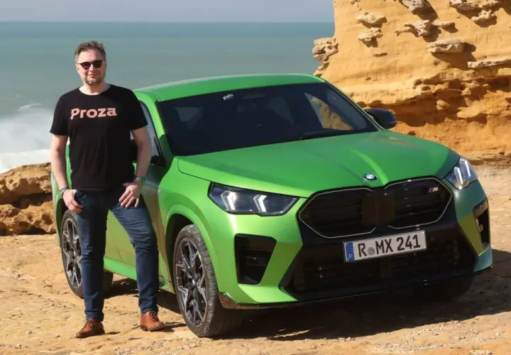 Mężczyzna w czarnej koszulce "Proza" stoi obok jaskrawozielonego BMW X2 M. Samochód zaparkowany jest na skalistym wybrzeżu z widokiem na wzburzone morze.