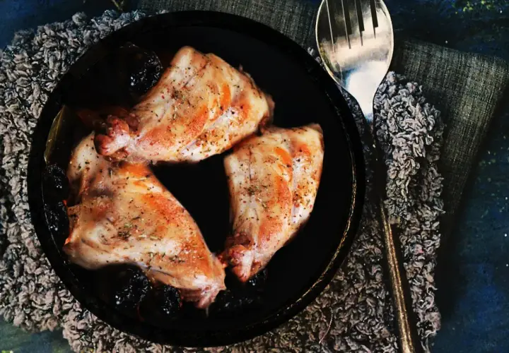 Trois morceaux de lapin aux pruneaux, assaisonnés et dorés, servis dans un plat noir avec une cuillère argentée.