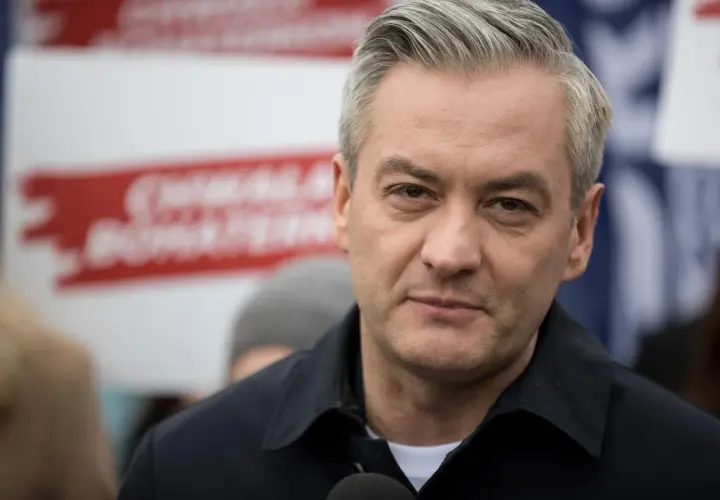 Robert Biedroń, polityk, w czarnej kurtce, z siwymi włosami. W tle czerwone napisy.