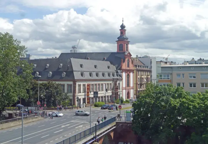 Barocke Kirche mit Turm und Mansarddach, Teil des Jakobsweg Hessen. Autos fahren auf einer Straße, Menschen überqueren sie.