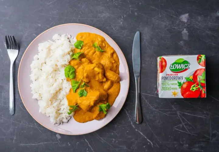Pyszny butter chicken przepis na talerzu z ryżem, udekorowany świeżą kolendrą. Obok nóż, widelec i przecier pomidorowy Łowicz.