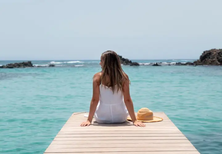 Mujer con vestido blanco sentada en un muelle de madera, disfrutando de la vista al mar turquesa. Su sombrero de paja descansa a su lado, perfecta para viajar sola.