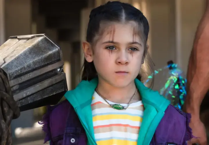 La fille de Thor, avec des marques sur le visage, porte une veste violette et un collier vert. À côté d'elle, le marteau Mjolnir.