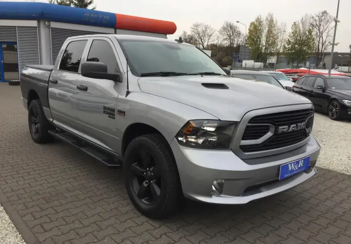 Srebrny Dodge Ram 1500 5.7 Hemi z czarnymi felgami i przyciemnianymi szybami stoi na parkingu przed budynkiem z niebiesko-pomarańczowym banerem.