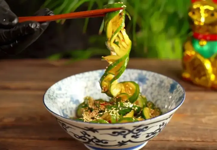 Chopsticks podnoszą zwinięty plasterek ogórka z miski pełnej tajskiej sałatki z ogórka, posypanej sezamem.