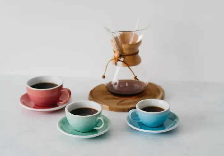 Chemex vs drip: trzy kolorowe filiżanki z kawą i szklany dzbanek Chemex na drewnianej podstawce.