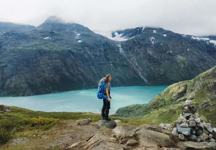 A hiker with a blue backpack stands on a rocky outcrop, overlooking a stunning turquoise lake in Jotunheimen National Park. The dramatic mountains behind are shrouded in mist.