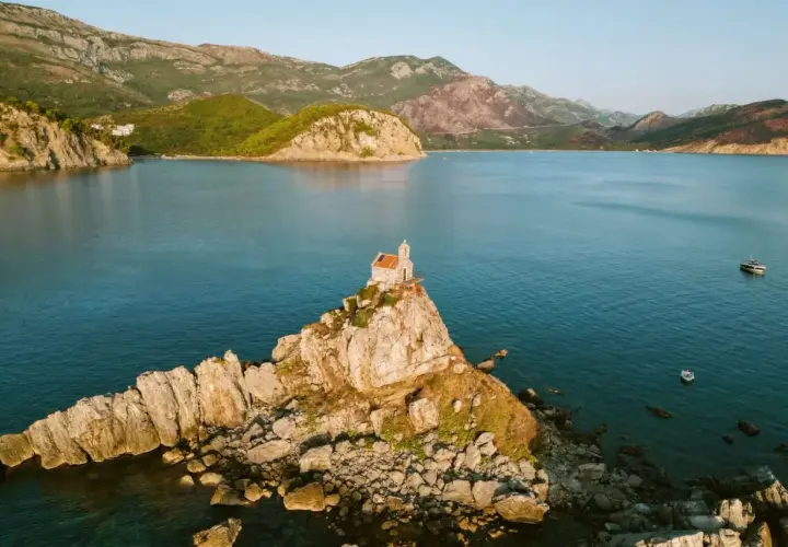 A tiny church perches on a rocky islet, a picturesque stop on a Montenegro itinerary. Mountains rise behind the tranquil blue bay.