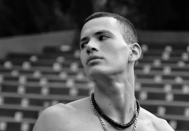 Joven con buzz cut largo, collar plateado y mirada perdida al horizonte.