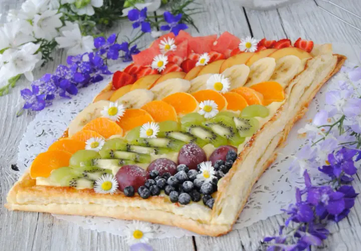 Kolorowa tarta z owocami: kiwi, mandarynki, banany, truskawki, winogrona i borówki, ozdobiona stokrotkami.