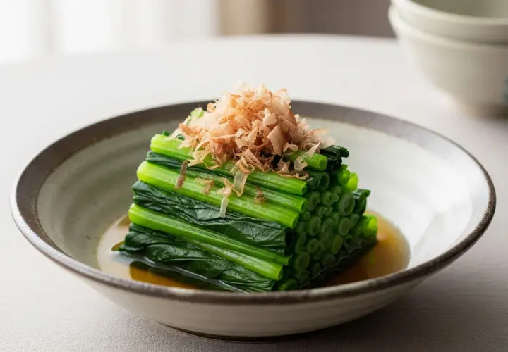 A neat stack of blanched spinach, known as spinach ohitashi, is served in a shallow bowl with a light broth and topped with delicate bonito flakes.
