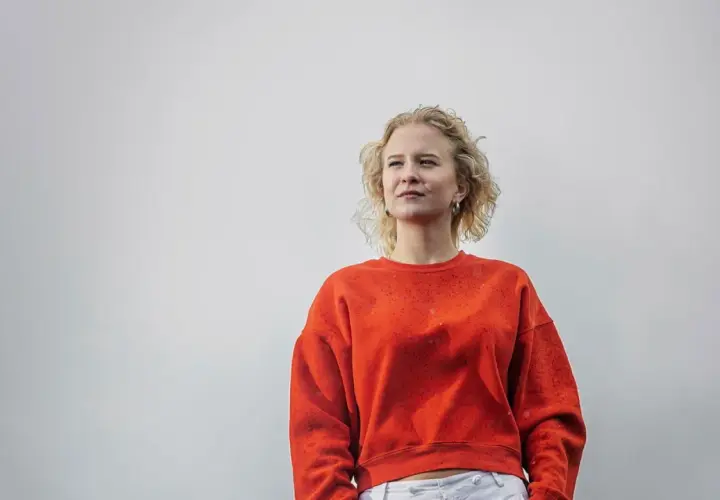 Sigrid Johnson, med sitt blonda, lockiga hår, bär en orange tröja och tittar drömmande bort.