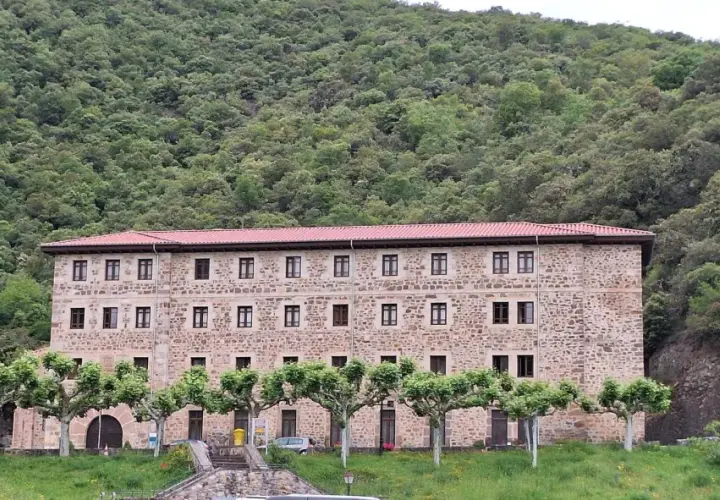Edificio de piedra con tejado rojo, rodeado de árboles y un monte frondoso. Es el monasterio de Santo Toribio de Liébana.