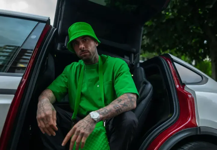 Malik Montana w zielonym kapeluszu i bluzie siedzi w otwartym samochodzie. Jego tatuaże i zegarek przyciągają wzrok, a jego wzrost jest imponujący.