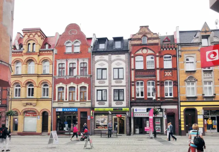 Kolorowe kamienice na lęborkim starym mieście, z witrynami sklepów i przechodniami na brukowanym placu.