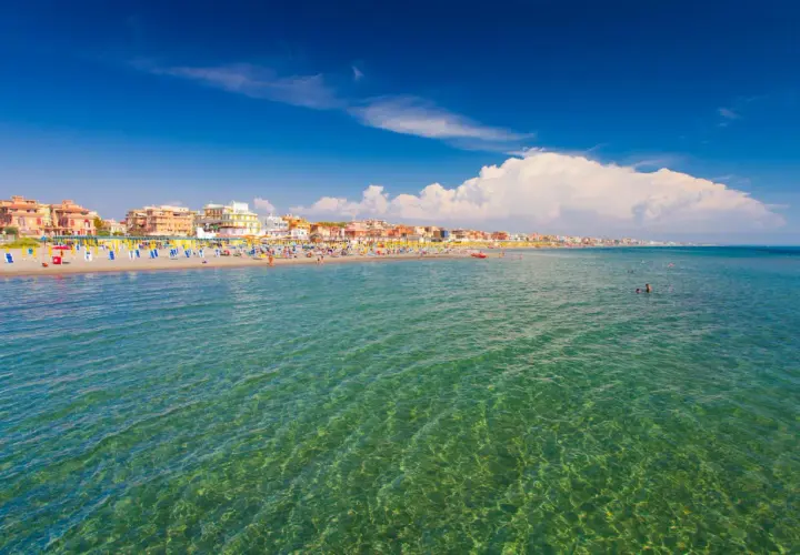 Słoneczny dzień nad morzem w Rzymie. Tłumy plażowiczów, kolorowe parasole i budynki wzdłuż wybrzeża.