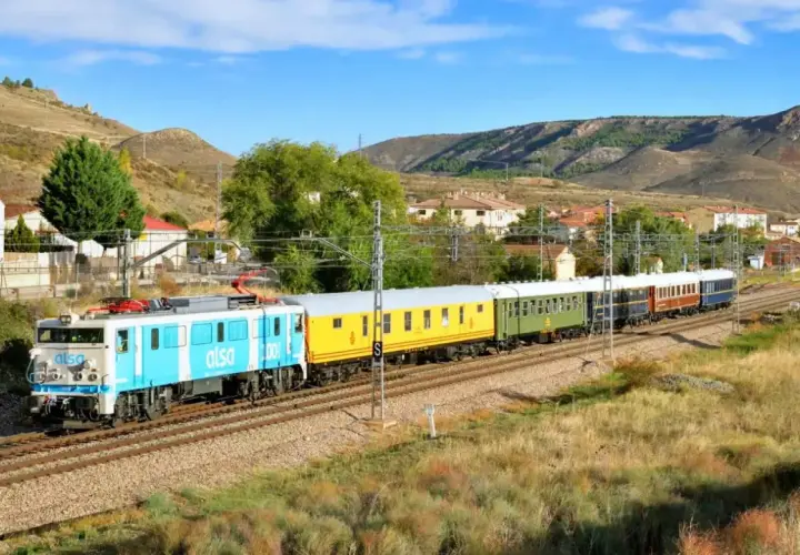 Un tren de Alsa, con vagones de colores, viaja por un paisaje montañoso. Este tren zaragoza logroño atraviesa un pueblo con casas blancas y tejados rojos.