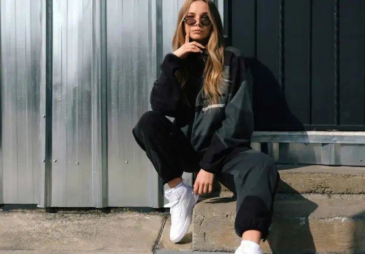 Chica con estilos de ropa mujer modernos, posa con sudadera y pantalones deportivos, zapatillas blancas y gafas de sol.