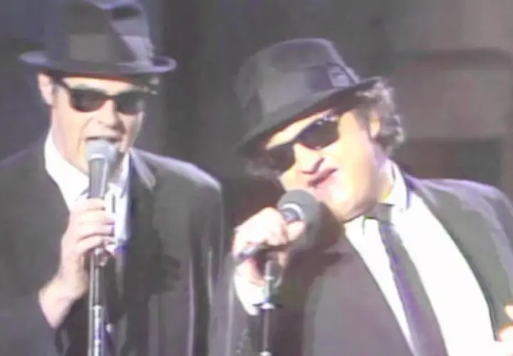 Dan Aykroyd giovane e John Belushi sul palco, entrambi con cappello e occhiali da sole, cantano al microfono.