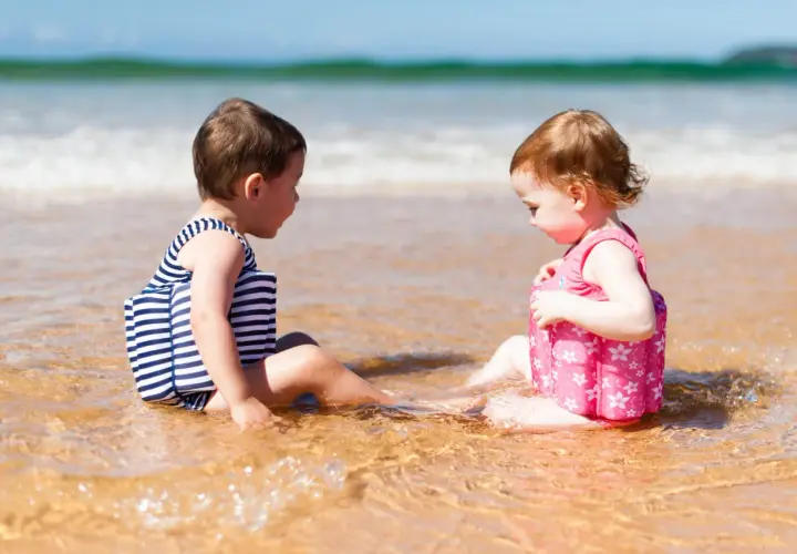 Dwoje dzieci w kostiumach kąpielowych z kamizelkami pływającymi siedzi na plaży.