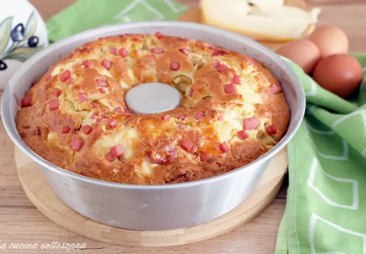 Gâteau salé végétarien doré, parsemé de dés de jambon et de fromage, dans un moule à savarin. Des œufs et du fromage sont visibles en arrière-plan.