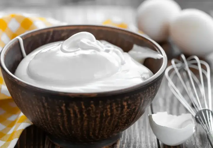 Un bol rempli de crème fouettée, blanc en neige, prêt à être utilisé pour une pâtisserie. Des œufs et un fouet sont visibles à proximité.