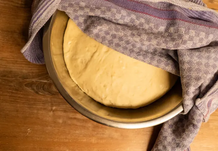 Ein goldgelber Pizzateig ruht in einer Schüssel, bedeckt mit einem karierten Tuch. Seine weiche, elastische pizzateig konsistenz verspricht eine köstliche Pizza.