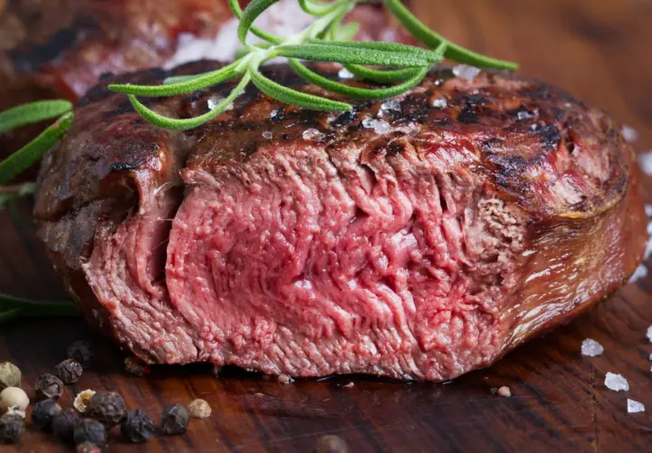 Succulenta tagliata di manzo al sangue, guarnita con rosmarino fresco e grani di pepe su un tagliere di legno.