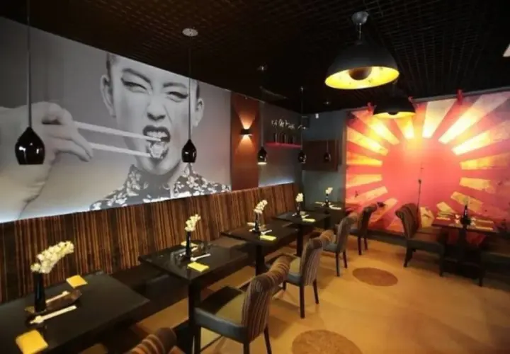 Szukasz miejsca, gdzie zjeść w Szczecinie? Wnętrze restauracji z azjatyckim klimatem, wielkoformatowym zdjęciem kobiety jedzącej pałeczkami i grafiką wschodzącego słońca.