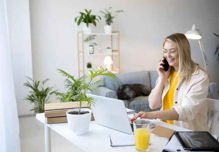 Uśmiechnięta kobieta pracuje przy laptopie i rozmawia przez telefon w domowym biurze.