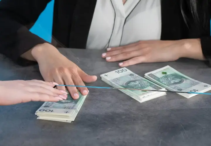 Dwie osoby przy blacie, jedna przekazuje drugiej stos banknotów 100 zł. Czy bank może żądać dowodu osobistego przy wpłacie?