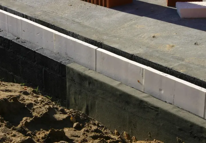 Biały styropian na fundamenty, izolujący czarną ścianę fundamentową od gruntu.