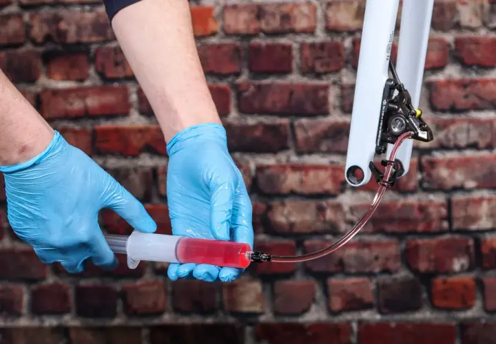 Hands in blue gloves bleed bike brakes, using a syringe filled with red fluid. This shows how much brake fluid to bleed brakes for optimal performance.