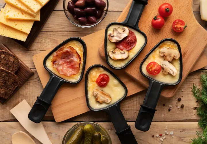 Raclette party with melted cheese, bacon, mushrooms, and tomatoes. Discover par quoi remplacer le fromage à raclette pour varier les plaisirs !