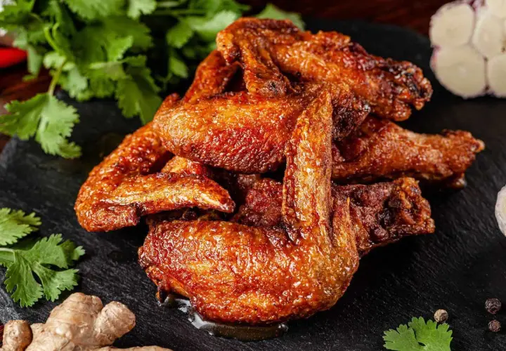 Alette di pollo al forno dorate e succulente, servite su una pietra scura con coriandolo fresco, zenzero e aglio.
