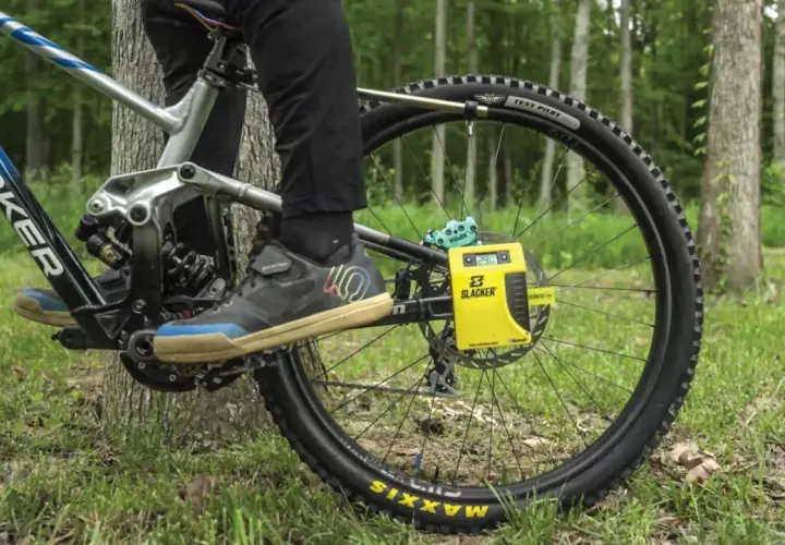 A person uses a yellow Slacker digital scale for rear shock measurements on a mountain bike, with a forest background.