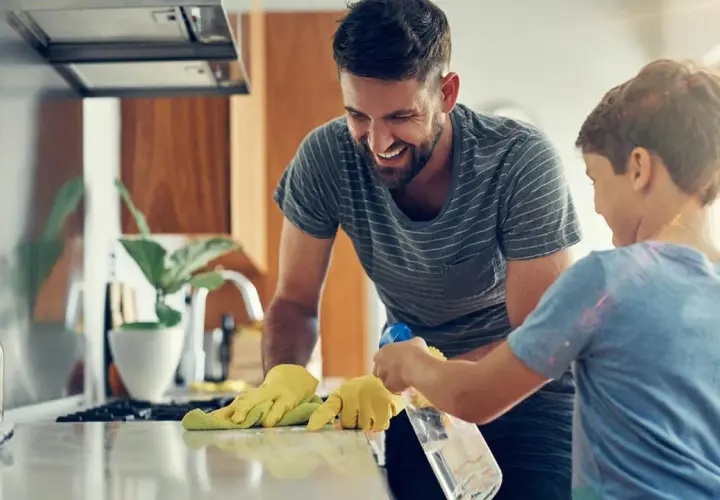 Padre e hijo ríen mientras aprenden como limpiar tu casa. El niño rocía la encimera mientras el padre la limpia con un paño.