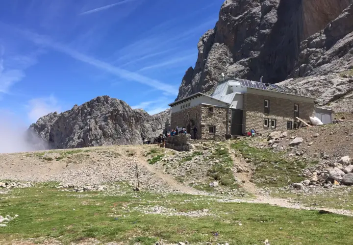 Schronisko pod masywnym **Naranjo de Bulnes**. Grupa turystów odpoczywa na tle skalistych szczytów i błękitnego nieba.