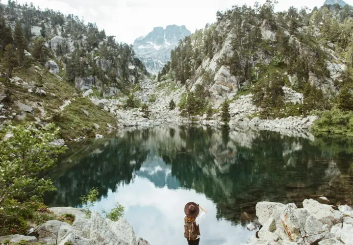 Una excursionista admira un lago sereno en el **parque nacional de los Pirineos**, rodeado de montañas rocosas y pinos.
