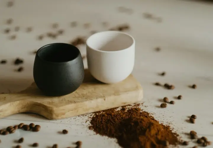Dwa ceramiczne kubki, czarny i biały, na drewnianej desce. Obok rozsypana mielona kawa, która sugeruje, czy kawe sypana sie miesza.