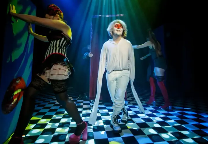 Scena teatralna, gdzie młody mężczyzna w białym stroju i makijażu twarzy stoi pośród postaci w kostiumach, tworząc atmosferę tragifarsy.