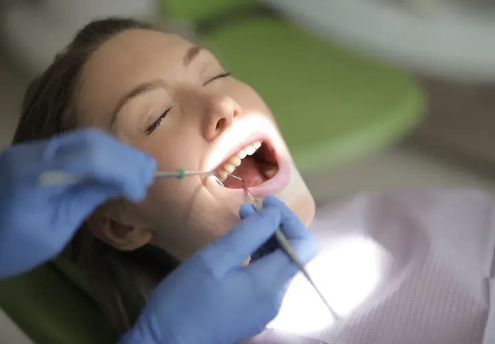 Kobieta z zamkniętymi oczami poddaje się zabiegowi stomatologicznemu. Dentysta w niebieskich rękawiczkach wykonuje kiretaż zamknięty, używając narzędzi do oczyszczania zębów.