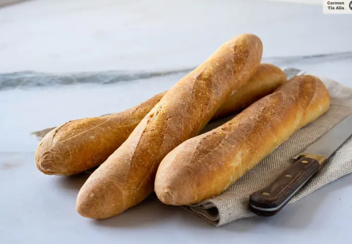 Tres baguettes doradas recién horneadas, listas para aprender como hacer baguette. Un cuchillo de madera descansa a su lado.