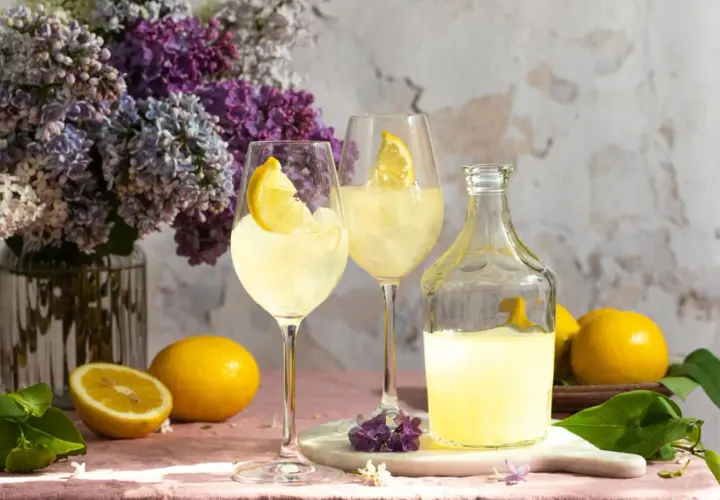 Erfrischendes Lemon Spritz Rezept: Zwei Gläser mit Eis und Zitronenscheiben, daneben eine Karaffe mit Zitronenlikör und frische Zitronen. Ein Blumenstrauß aus Flieder im Hintergrund.