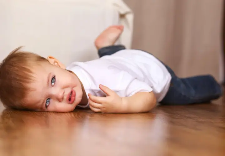 Un bebé de 16 meses con ojos azules y cabello castaño yace en el suelo de madera, vestido con una camiseta blanca y jeans.
