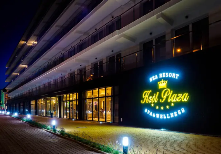 Nocny widok na nowoczesny hotel Król Plaza Spa & Wellness w Jarosławcu. Neonowy napis zachęca do sprawdzenia opinii o tym miejscu.