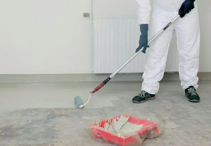 Person i skyddsdräkt målar betonggolv med roller. En färgtråg med ljus färg står på golvet.