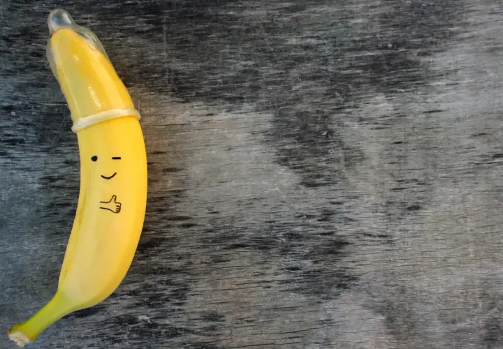 Banan z namalowaną buźką i kciukiem w górę, ubrany w **kondom**. To zabawne przypomnienie o bezpieczeństwie.