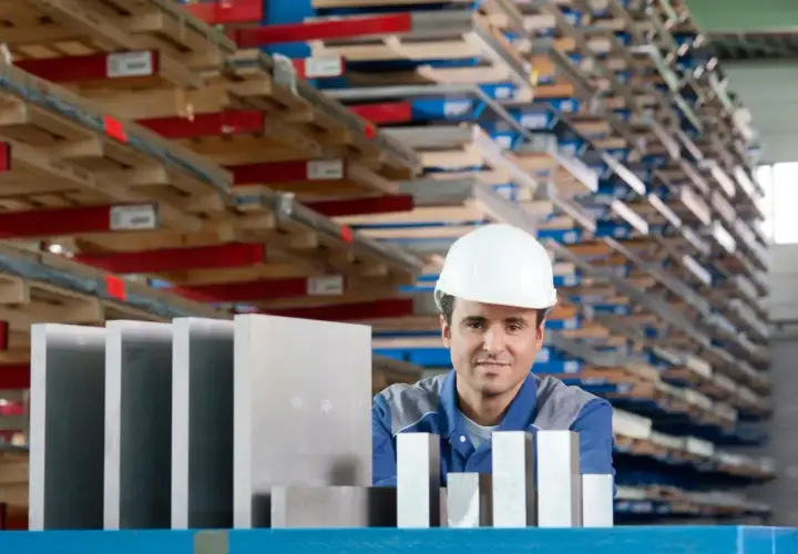 Pracownik w białym kasku stoi przed regałami pełnymi materiałów, obok niego leżą różne stopy aluminium.